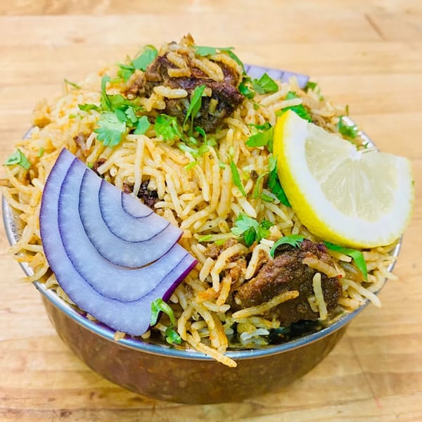 Grand India's lamb biryani with rice, cilantro, red onion, and a lemon wedge in a metal bowl.